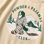 Sand colored sweatshirt with a dog in plaid pajamas and skis, powder and pajamas club text, and snowy landscape design.