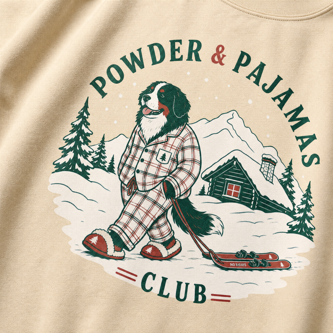 Sand colored sweatshirt with a dog in plaid pajamas and skis, powder and pajamas club text, and snowy landscape design.