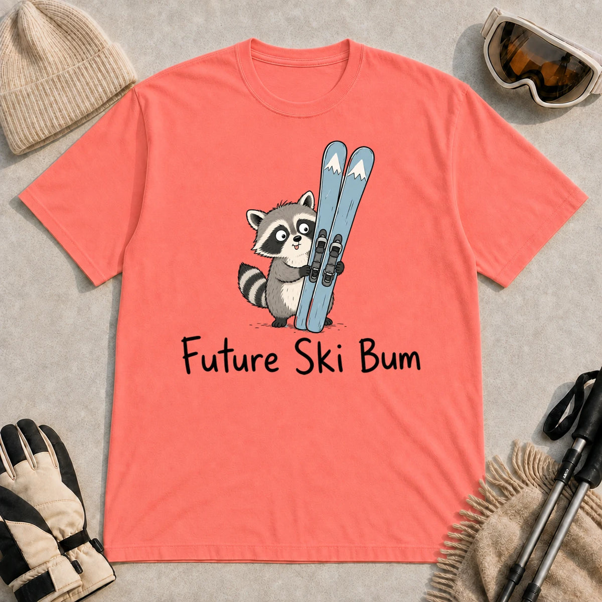 Coral Silk tee featuring a cute raccoon standing beside a pair of skis, with the phrase “Future Ski Bum.”