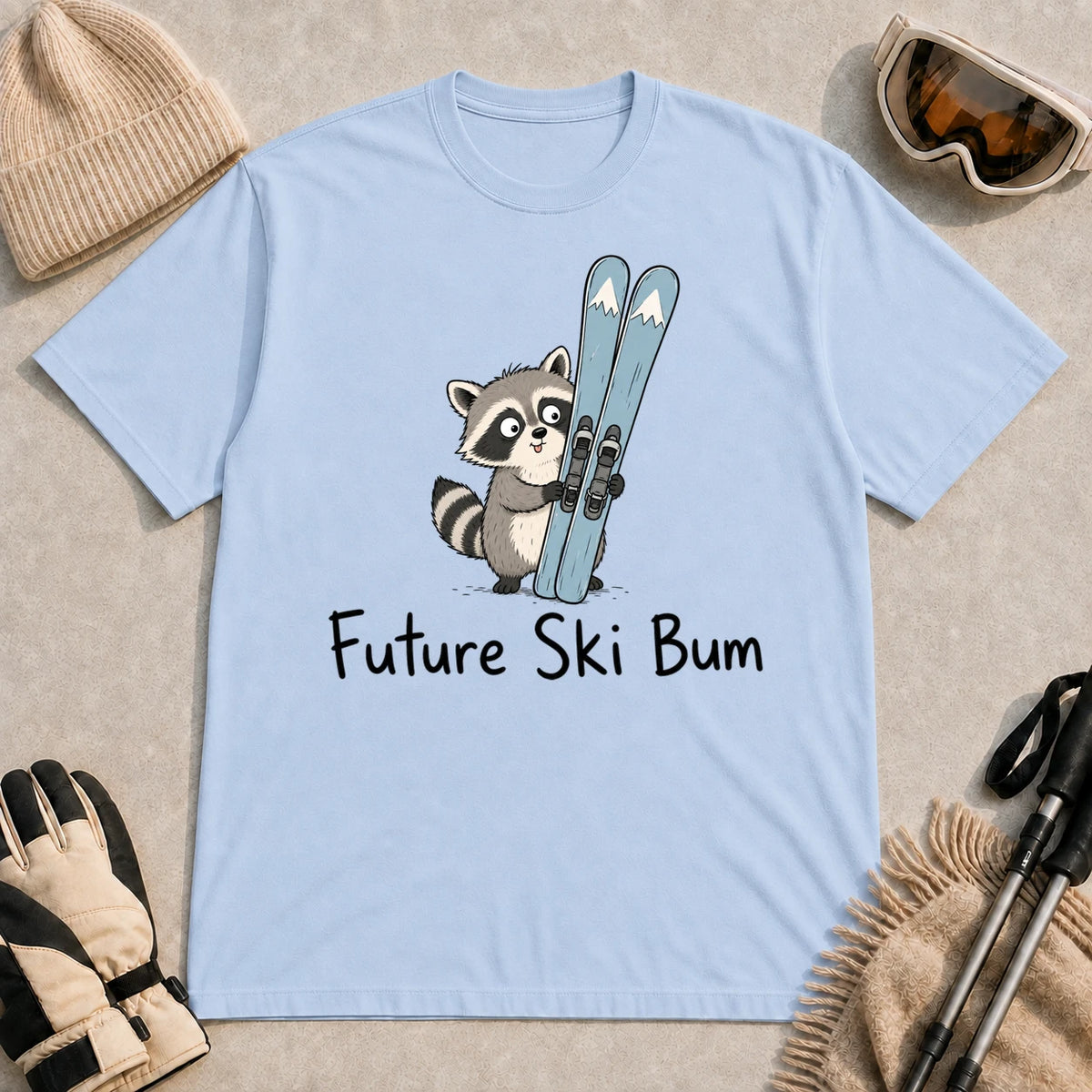 Light Blue tee featuring a cute raccoon standing beside a pair of skis, with the phrase “Future Ski Bum.”