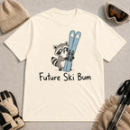 Natural tee featuring a cute raccoon standing beside a pair of skis, with the phrase “Future Ski Bum.”