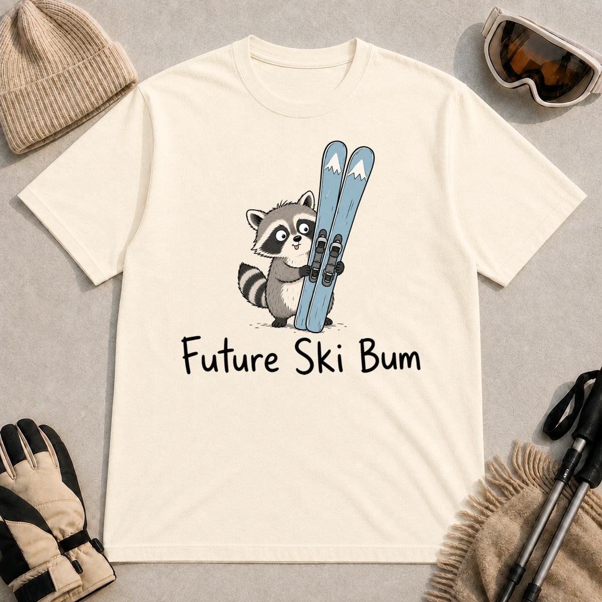 Natural tee featuring a cute raccoon standing beside a pair of skis, with the phrase “Future Ski Bum.”