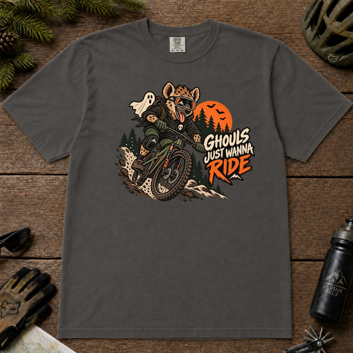 Pepper tee featuring a hyena mountain biking through a spooky Halloween trail scene with a ghost, bats, and orange moon beside the phrase “Ghouls Just Wanna Ride.”