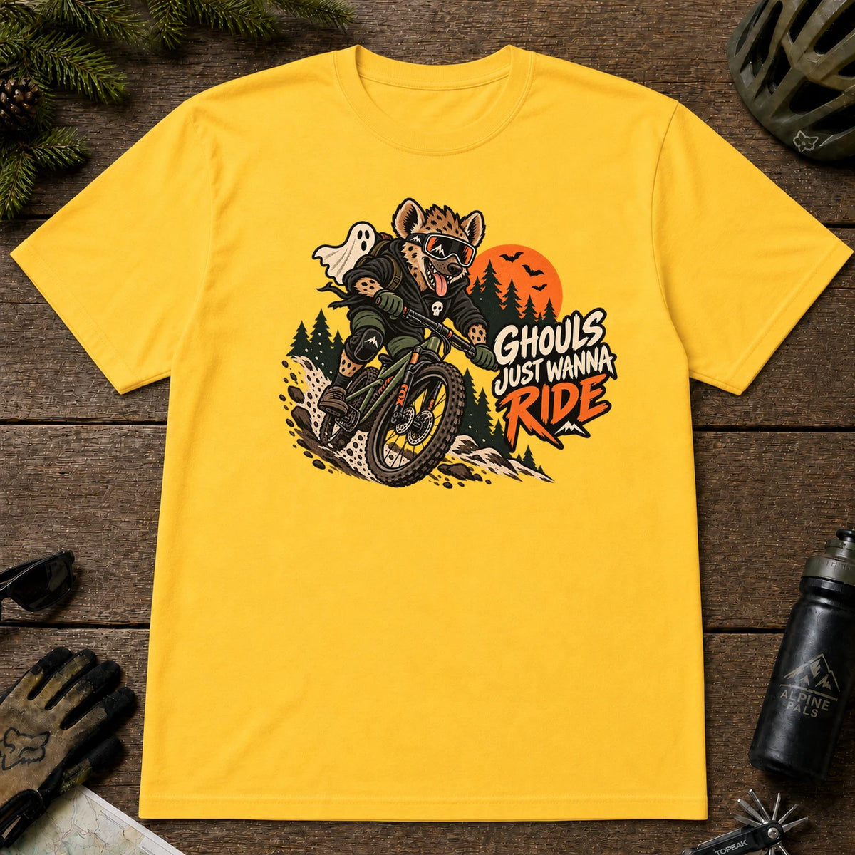 Daisy tee featuring a hyena mountain biking through a spooky Halloween trail scene with a ghost, bats, and orange moon beside the phrase “Ghouls Just Wanna Ride.”