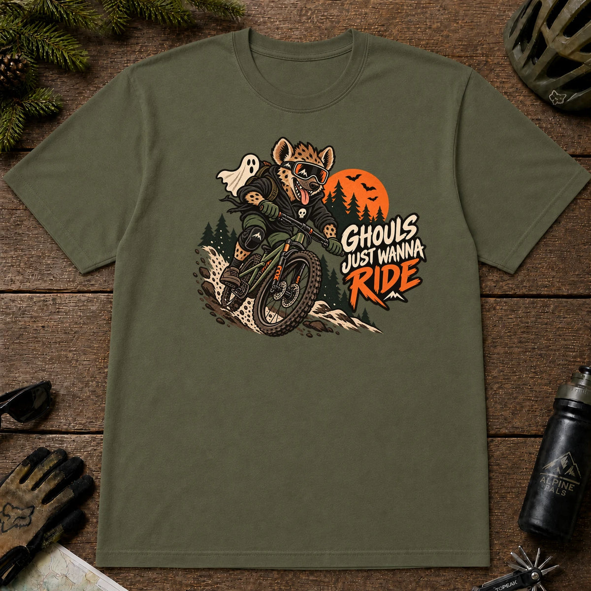 Military Green tee featuring a hyena mountain biking through a spooky Halloween trail scene with a ghost, bats, and orange moon beside the phrase “Ghouls Just Wanna Ride.”