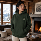 Woman wearing a forest green hoodie with a logo in a cozy living room with a fireplace and mountain view.