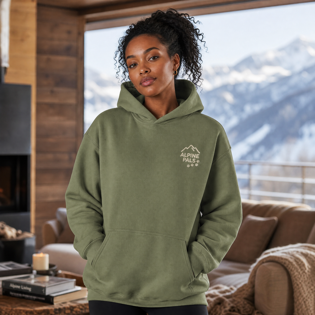 Woman wearing a military green hoodie with 'Alpine Pals' logo in a cozy living room with mountain view.