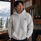 Man wearing an ash gray hoodie with a logo, standing in a cozy indoor setting with mountains in the background.