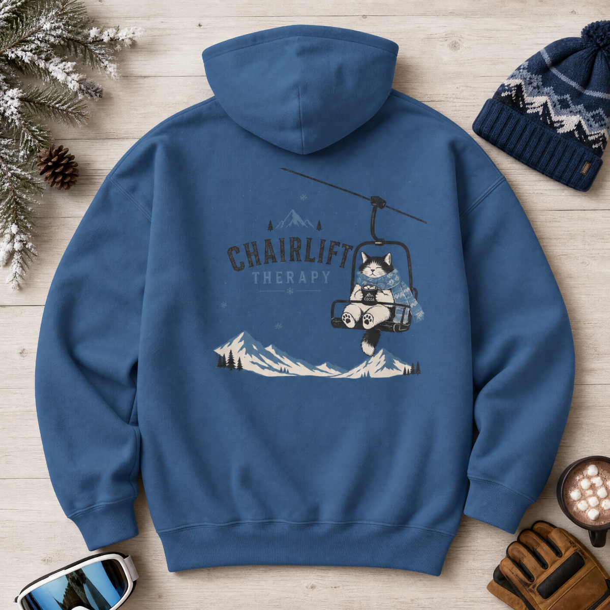 Indigo blue hoodie with a chairlift graphic on a wooden surface with winter accessories.