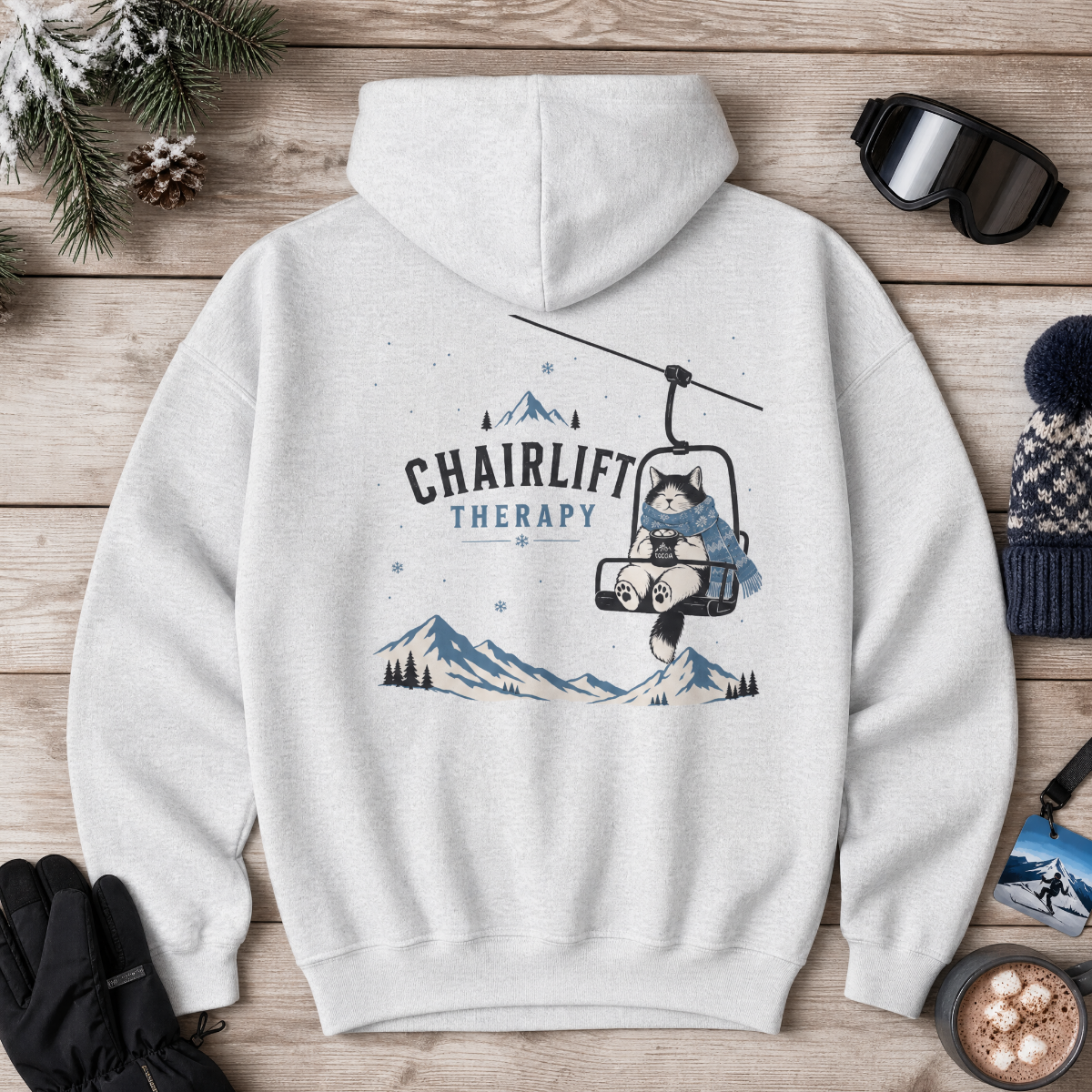 Ash gray hoodie with 'Chairlift Therapy' graphic on a wooden surface with winter accessories.