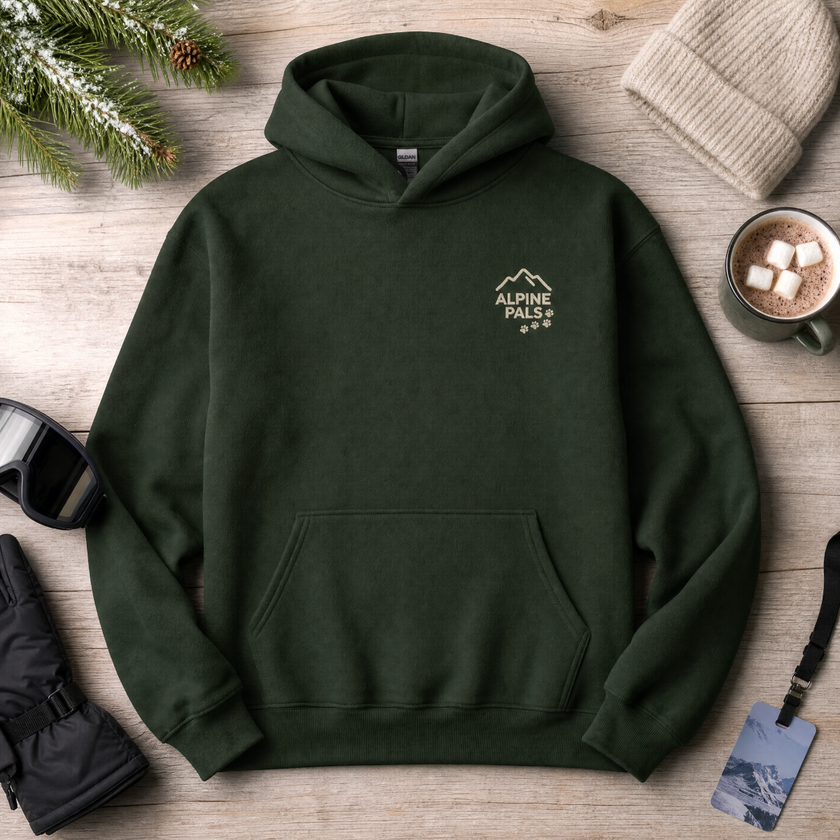 Forest green hoodie with 'Alpine Pals' logo on a wooden surface with winter accessories.