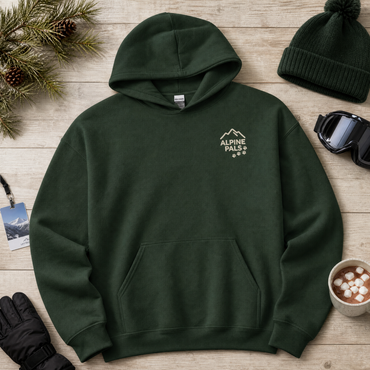 Forest green hoodie with 'Alpine Pals' logo on a wooden surface with winter accessories.