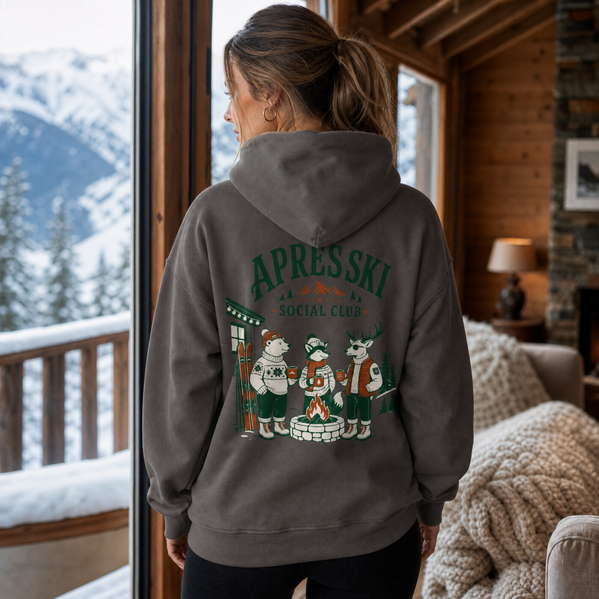 Person wearing a hoodie with 'Apres Ski Social Club' design in a cozy room with a snowy mountain view.