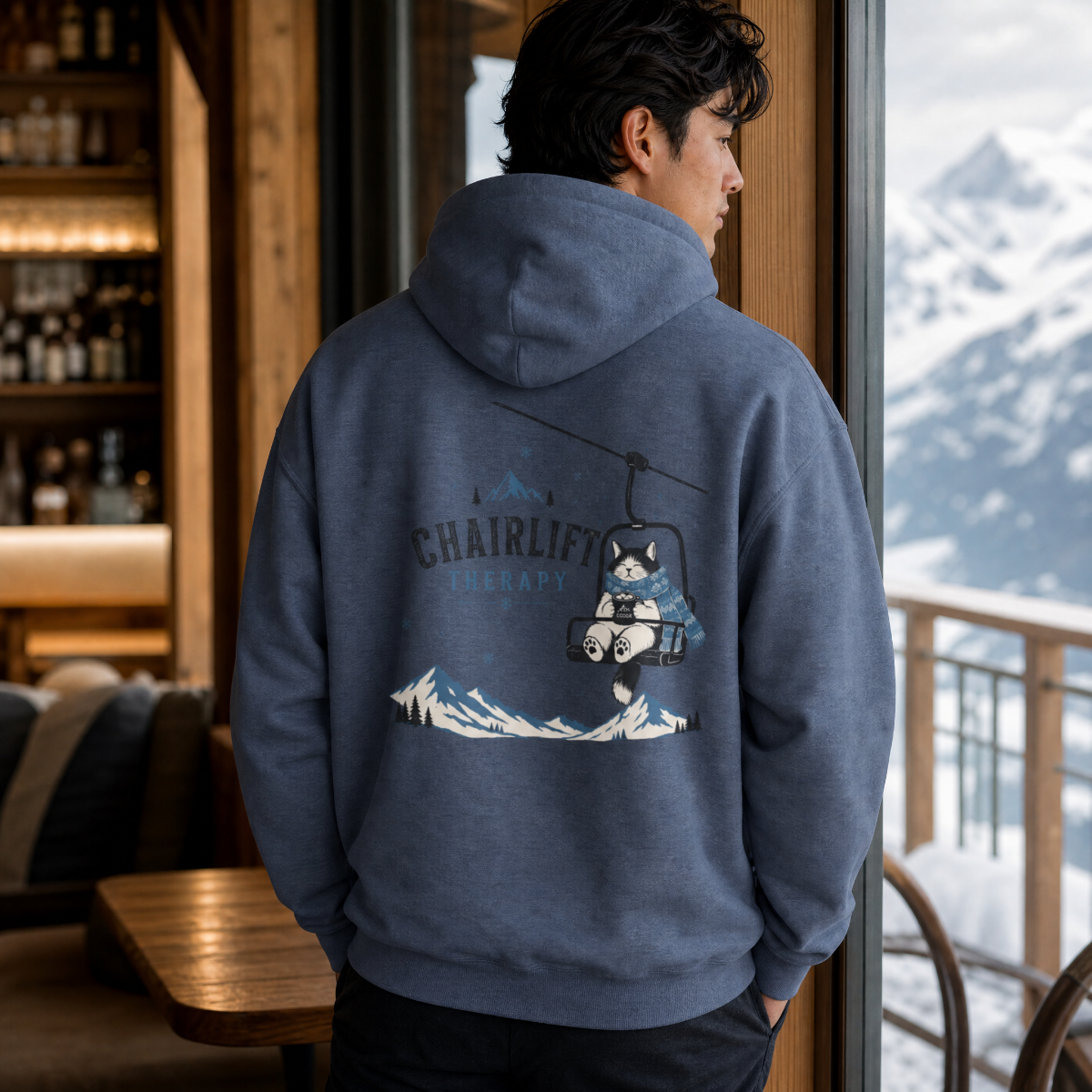 Person wearing a heather sport dark navy hoodie with a chairlift graphic, standing by a window with a mountain view.