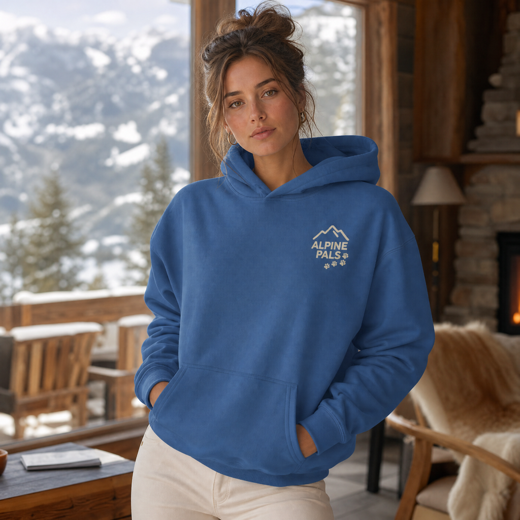 Woman wearing an indigo blue hoodie with 'Alpine Pals' logo in a cozy room with snowy mountain view.