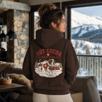Person wearing a 'Hot Cocoa Patrol' hoodie looking out at snowy mountains.
