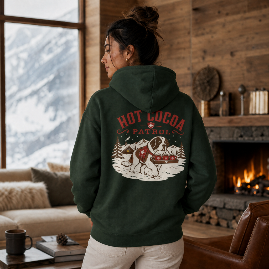 Person wearing a hoodie with 'Hot Cocoa Patrol' design in a cozy living room with a fireplace and mountain view.
