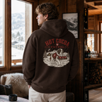 Person wearing a hoodie with 'Hot Cocoa Patrol' design, standing in a cozy room with a view of mountains.