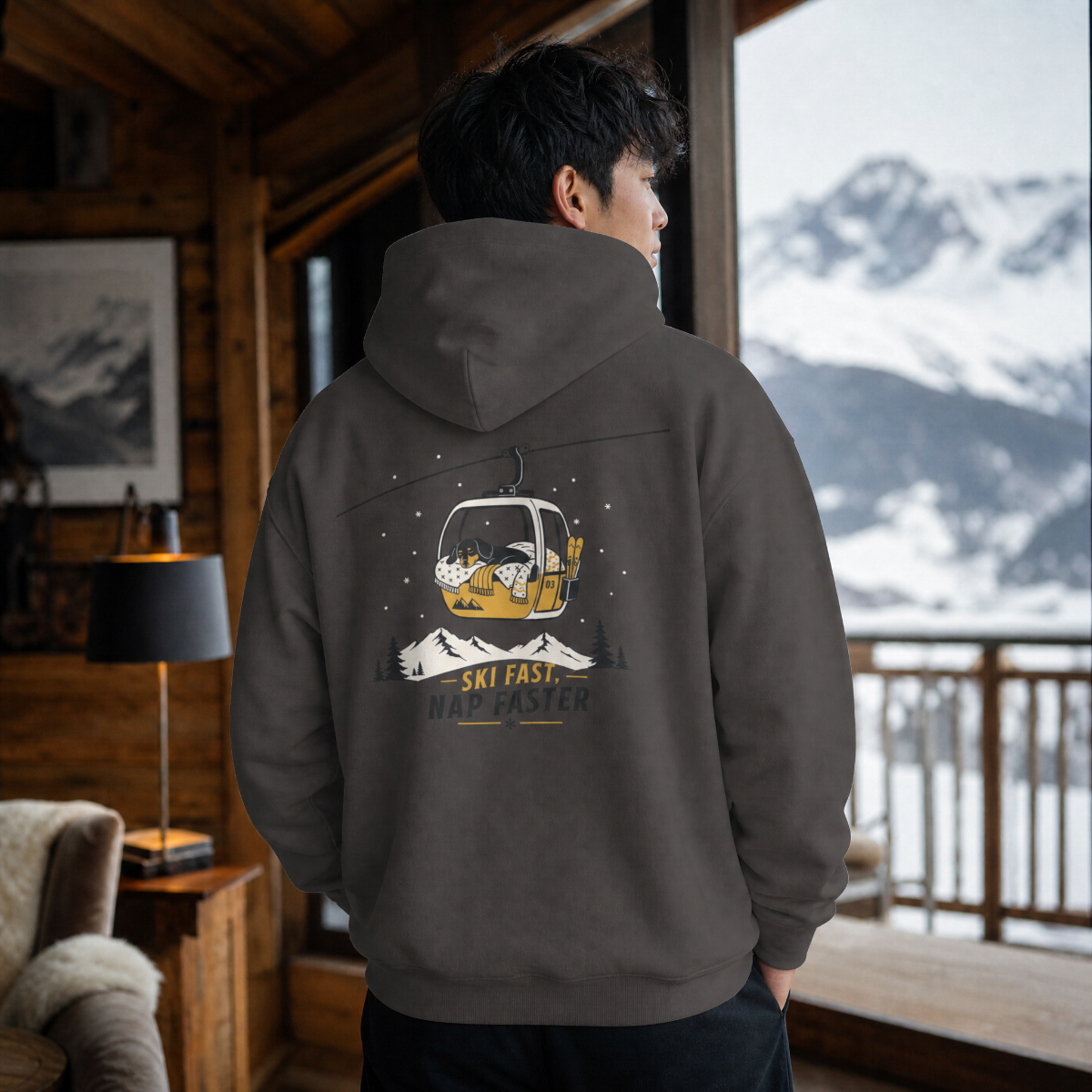 Person wearing a hoodie with a mountain graphic in a cozy room with a view of snow-covered mountains.