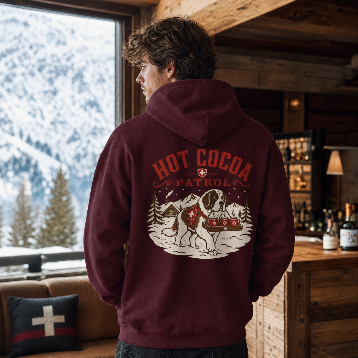 Person wearing a maroon hoodie with 'Hot Cocoa Patrol' design in a cozy indoor setting with snowy outdoors.