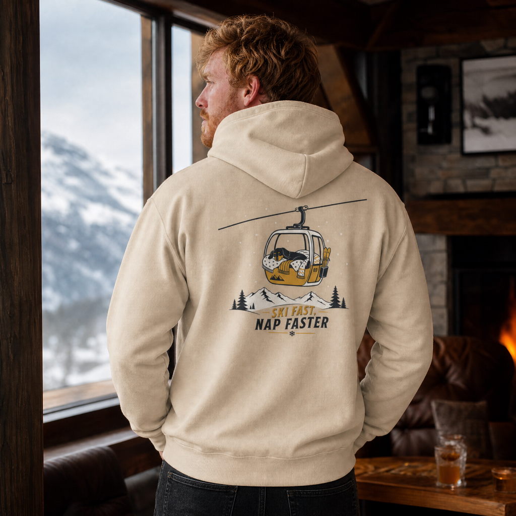 Man wearing a sand colored hoodie with a graphic design, standing in a cozy room with a view of mountains.