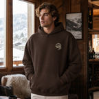 Man wearing a dark chocolate brown hoodie with 'Alpine Pals' logo in a cozy indoor setting with snowy landscape outside.