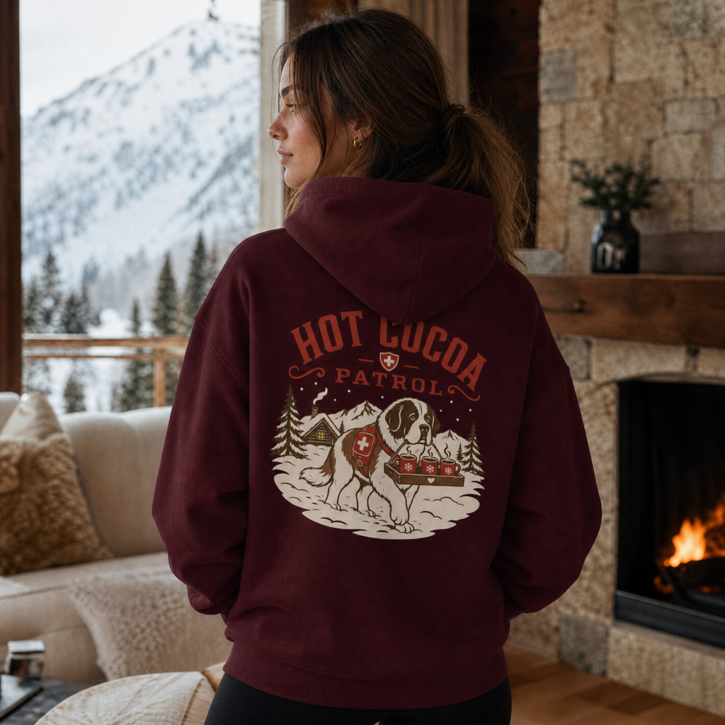 Person wearing a maroon hoodie with 'Hot Cocoa Patrol' design in a cozy living room with a fireplace and snowy mountain view.