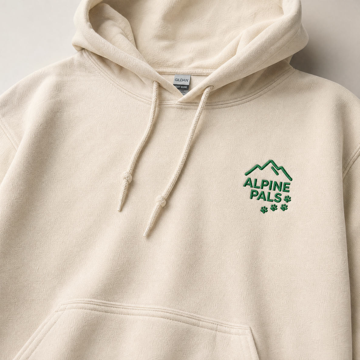 Sand colored hoodie with 'Alpine Pals' logo on a light gray background