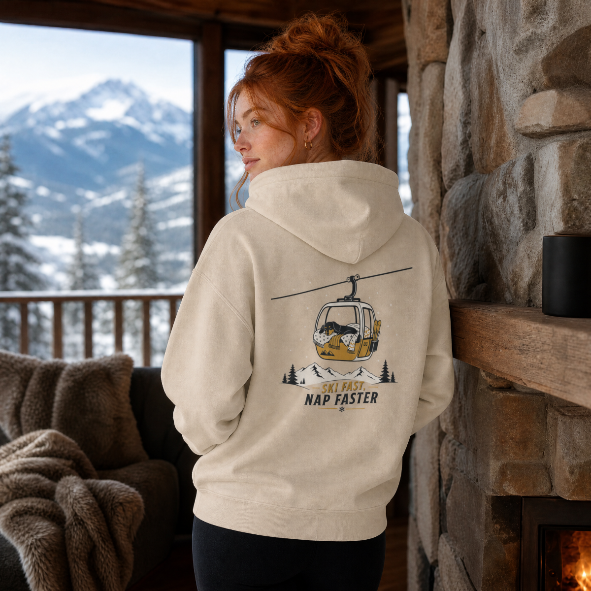 Person wearing a hoodie with a mountain graphic and text, standing in front of a stone fireplace with a snowy mountain view.