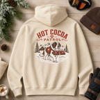 Sand colored hoodie with 'Hot Cocoa Patrol' design on a wooden surface with winter accessories.
