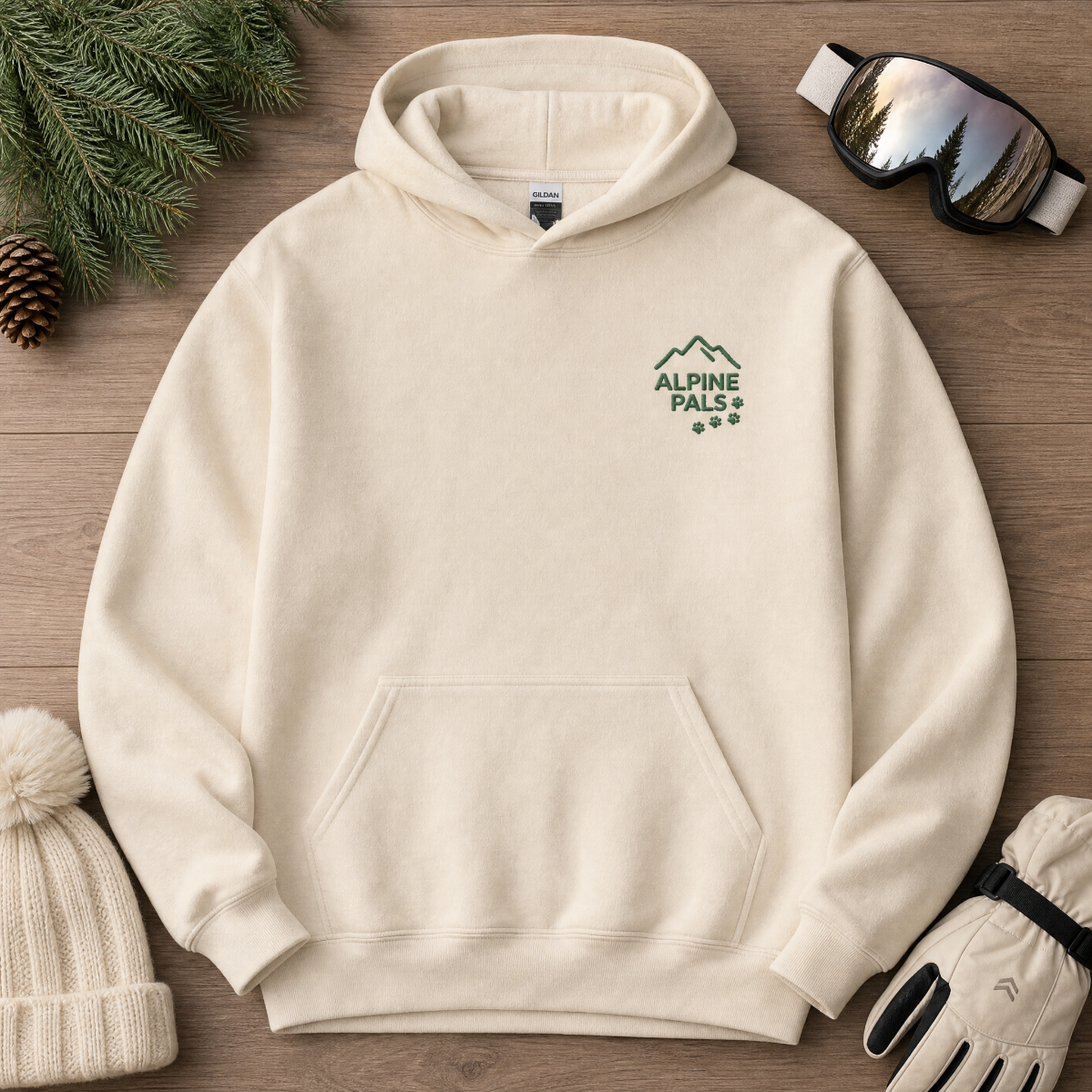 Sand colored hoodie with 'Alpine Pals' logo on a wooden surface with winter accessories.