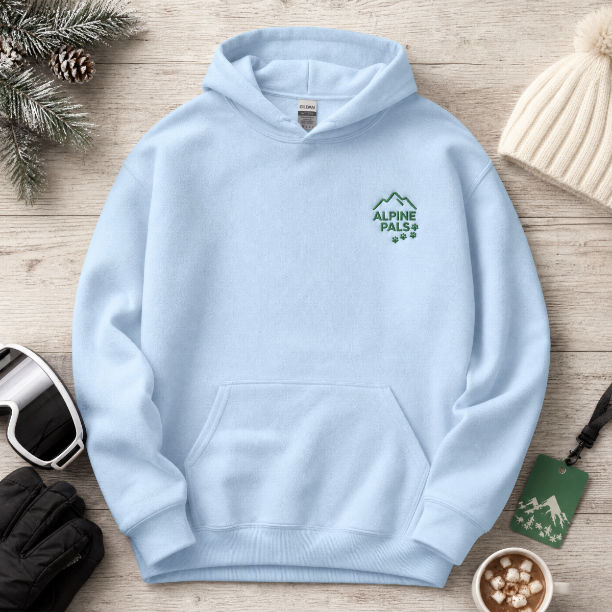 Light blue hoodie with 'Alpine Pals' logo on a wooden surface with winter accessories.