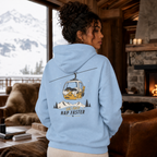 Person wearing a light blue hoodie with a mountain and cable car design, standing in a cozy living room with a snowy outdoor view.