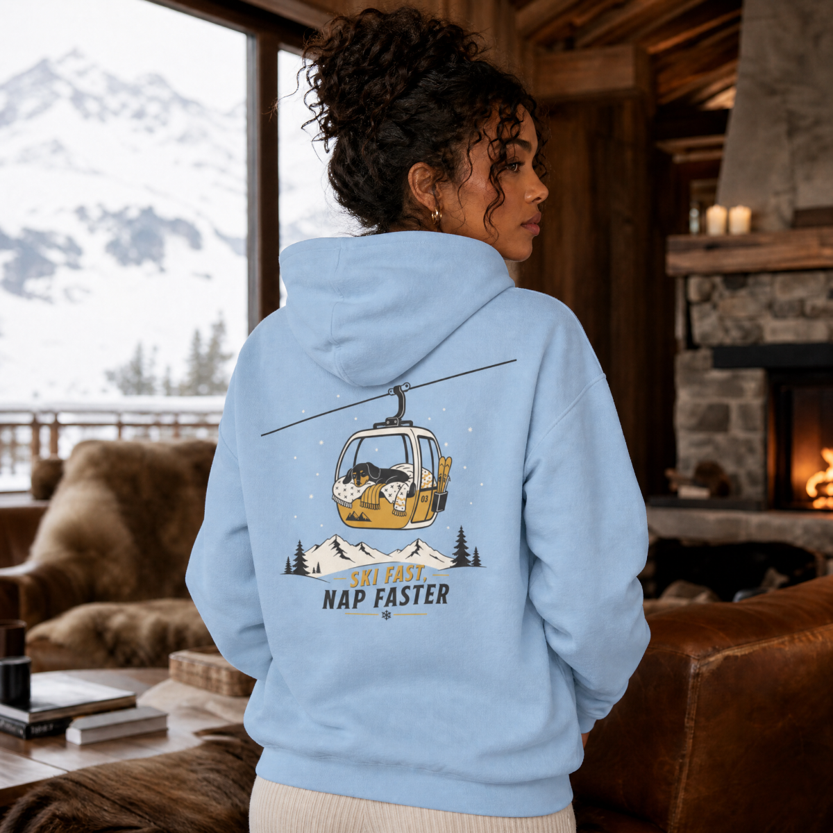 Person wearing a light blue hoodie with a mountain and cable car design, standing in a cozy living room with a snowy outdoor view.
