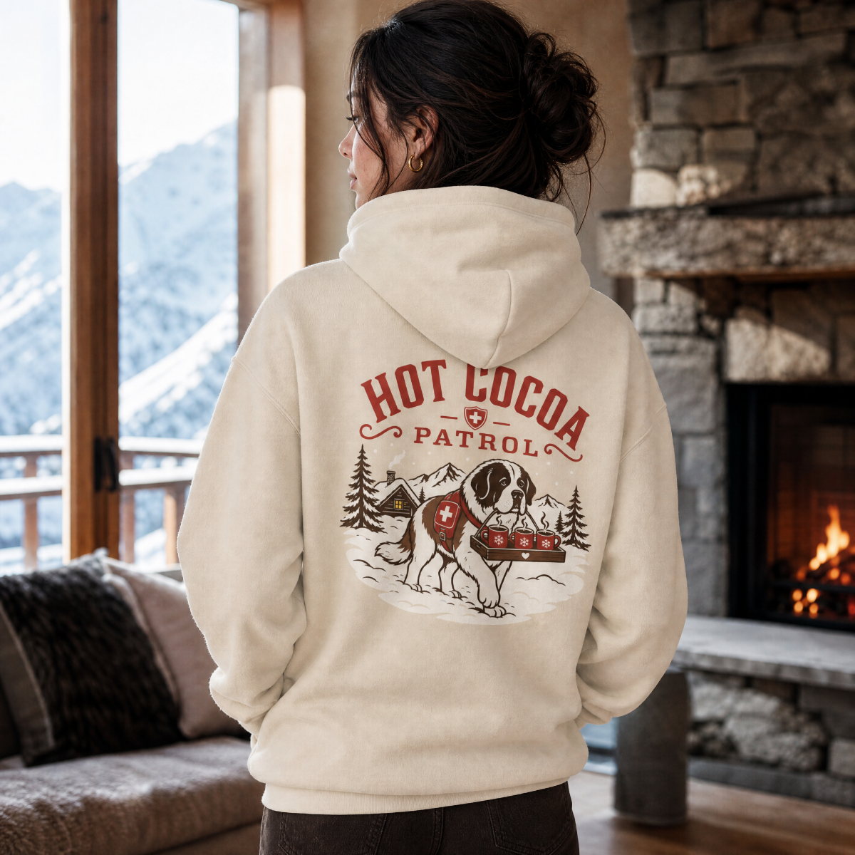 Person wearing a hoodie with 'Hot Cocoa Patrol' design in a cozy room with a fireplace and mountain view.