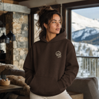 Woman wearing a dark chocolate brown hoodie with 'Alpine Pals' logo in a cozy indoor setting with mountain view.