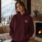 Woman wearing a maroon hoodie with 'Alpine Pals' logo in a cozy living room with a fireplace and snowy mountain view.