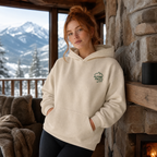 Woman wearing a sand colored hoodie with 'Alpine Pals' logo in a mountainous setting.