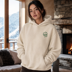 Woman wearing a sand colored hoodie with a logo in a cozy room with a fireplace and mountain view.