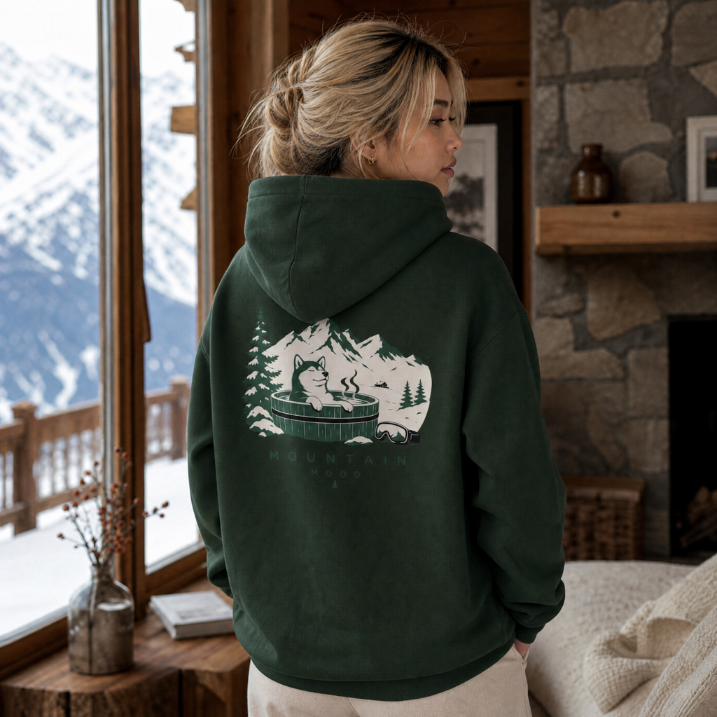 Person wearing a forest green hoodie with a mountain and bear design, standing in a cozy room with a snowy mountain view.