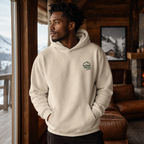 Man in a cozy cabin setting wearing a sand colored hoodie with the "Alpine Pals" logo.