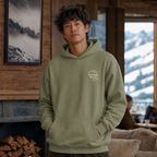 Man wearing a military green hoodie with a mountain logo in a cozy indoor setting with mountain views.