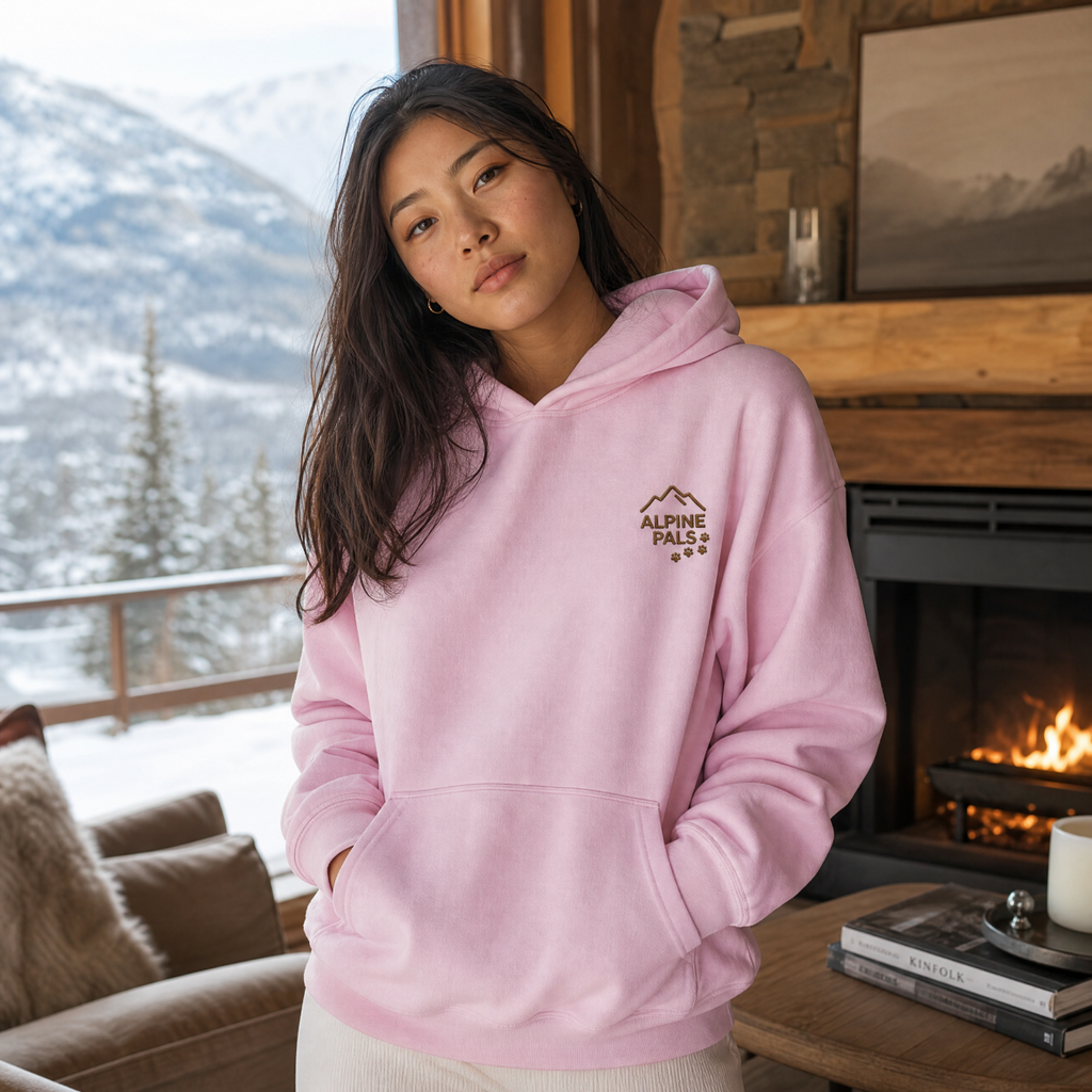 Person wearing a pink hoodie with 'Alpine Pals' logo in a cozy living room with a fireplace and mountain view.