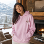 Person wearing a pink hoodie with 'Alpine Pals' logo in a cozy living room with a fireplace and mountain view.
