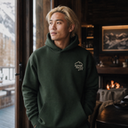 Man wearing a forest green hoodie with 'Alpine Pals' logo in a cozy indoor setting with a fireplace and mountain view.