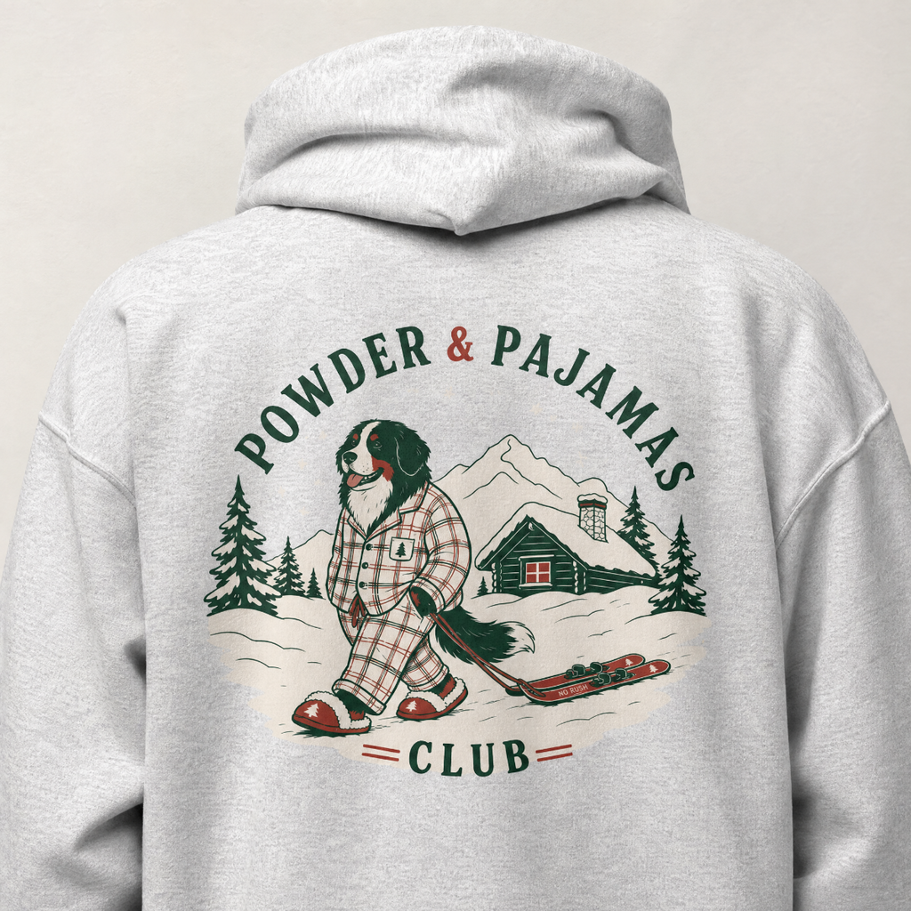 Ash gray hoodie with a graphic of a person in plaid pajamas skiing, text 'Powder & Pajamas Club', and a cabin in the mountains on a light gray background.