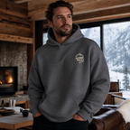 Man wearing a dark heather gray hoodie with a logo in a cozy indoor setting with a fireplace and snowy outdoor view.