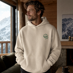 Man wearing a sand colored hoodie with a logo, standing in a cozy room with a mountain view.