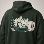 Forest green hoodie with a graphic of a dog in a hot tub and mountains on the back, on a light gray background.