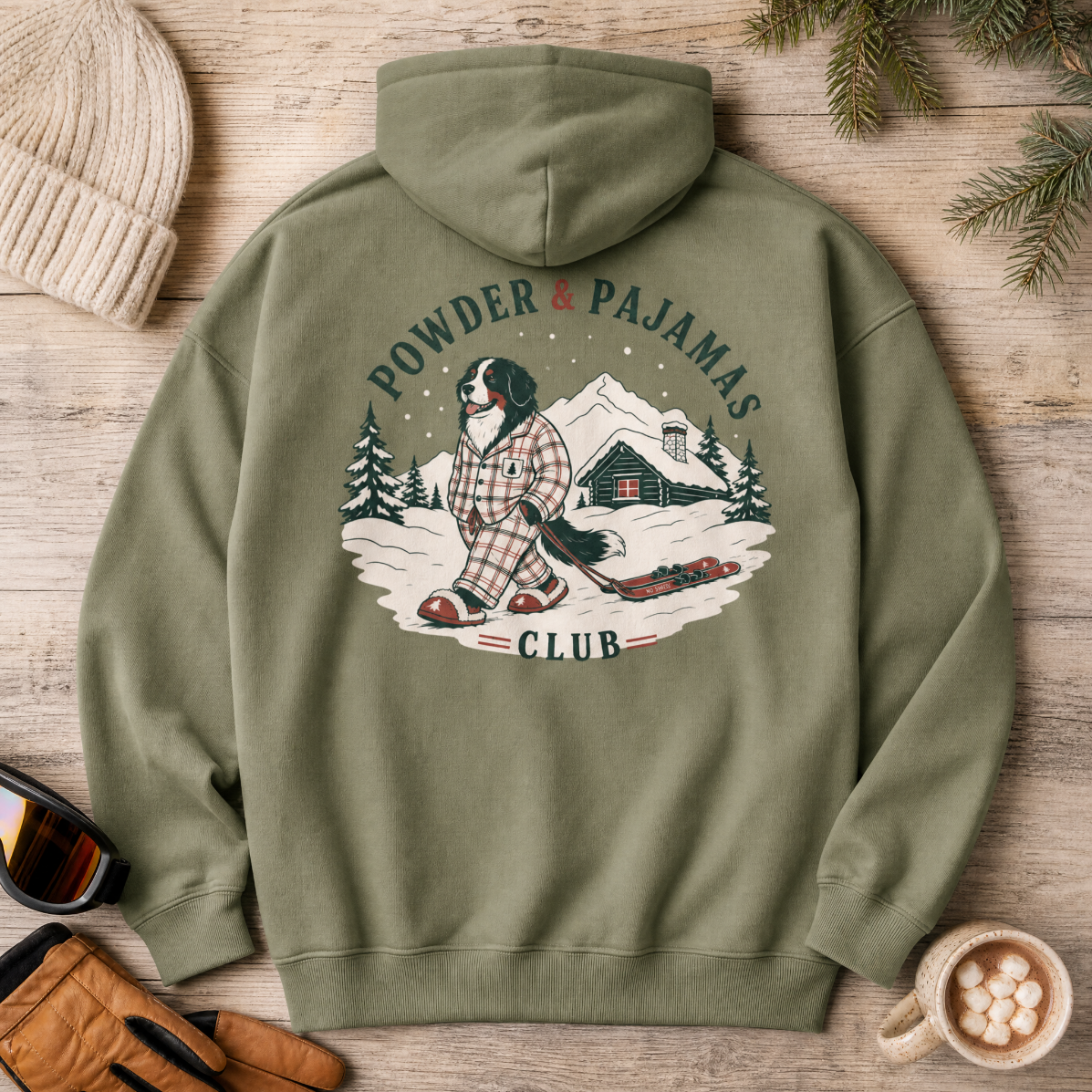 Military green hoodie with 'Powder & Pajamas Club' design on a wooden surface with winter accessories.